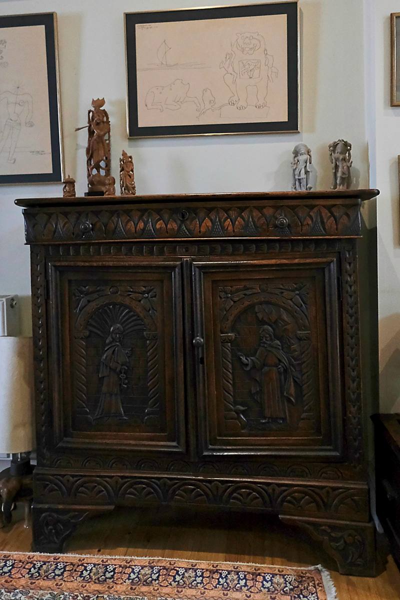 A carved dark wooden cabinet with small figures on top and a framed drawing above